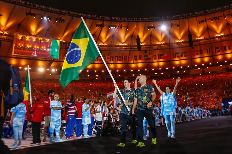 Ricardinho segura a bandeira do Brasil durante a entrada da delegação em um estádio lotado, à noite, ao lado de outros atletas.